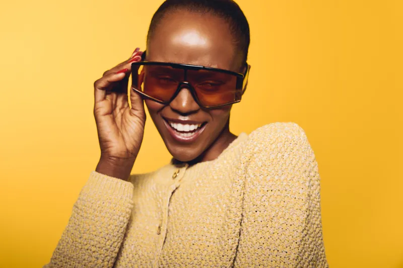 Smiling African woman with dark skin wearing oversized sunglasses and knitted sweater poses against yellow background. Concept for fashion, style, and joyful mood with vibrant colors.