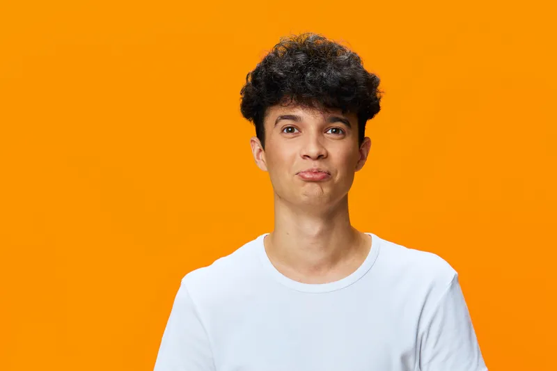 Young man with curly hair wearing white t-shirt posing with playful expression against bright orange background, casual style, studio portrait, vibrant colors.