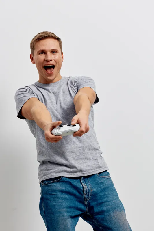 joyful man expressing excitement with a video game controller, casual grey t-shirt and jeans, standing against plain white background, lifestyle entertainment concept
