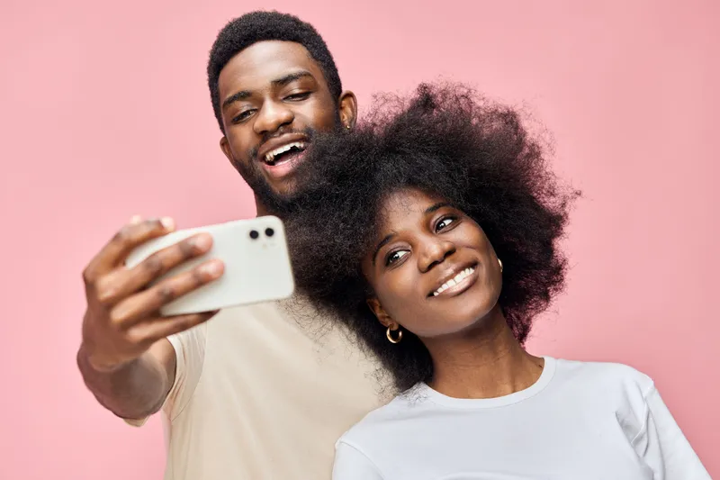 Young African couple taking a selfie against a pink background, expressing joy and love, showcasing vibrant clothing and a modern, playful style