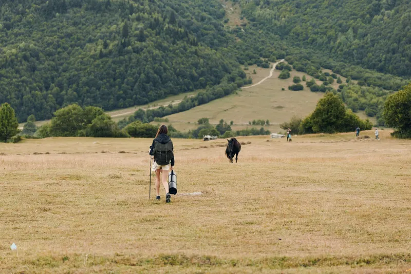 A lone traveler walks across a wide rural field toward distant hills, carrying a guitar case and backpack, capturing a quiet moment of outdoor exploration.