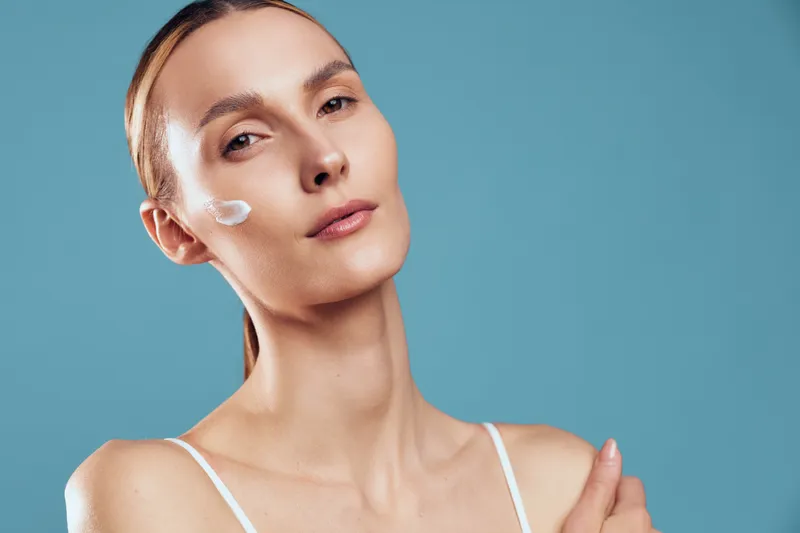 A young woman with clear skin applies moisturizing cream to her cheek against a blue background. The image represents skincare, beauty routine, and natural complexion care.