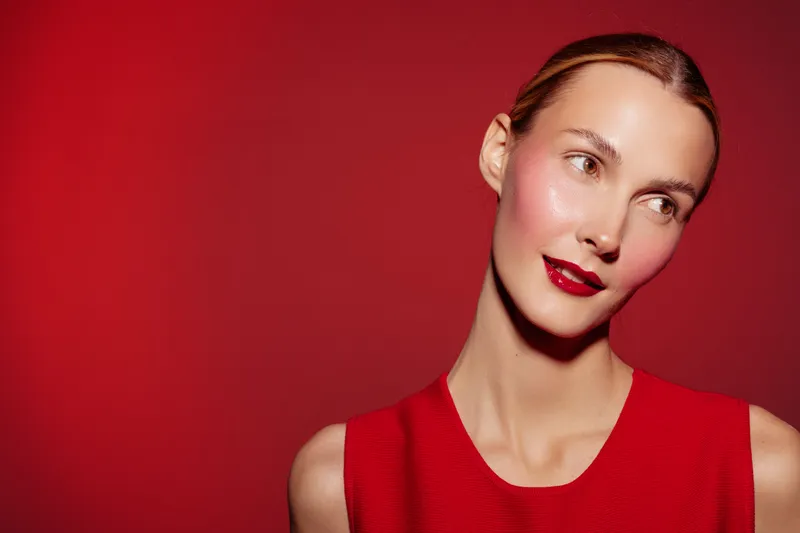 Young woman with shiny skin and red lipstick looks sideways on red background. Beauty portrait with smooth skin and elegant makeup. Concept of glamour and fashion.