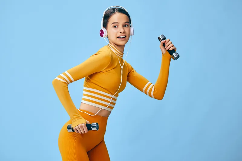 Woman fitness enthusiast in orange activewear lifts dumbbells with headphones against a blue backdrop, showcasing energy, movement and a dynamic workout moment.