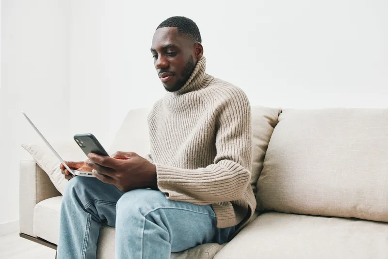 Young African man in cozy sweater using smartphone and laptop at home, focused on digital communication and online tasks, modern lifestyle, casual style