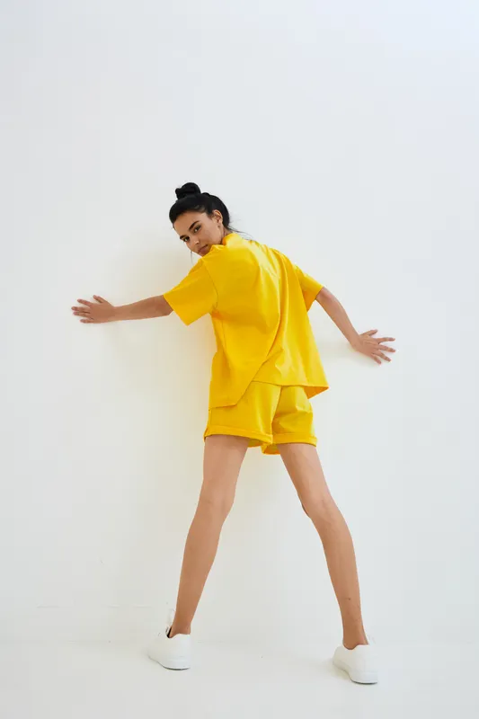 Young woman wearing bright yellow casual outfit and white sneakers stands against white wall, posing with arms stretched out, showing modern minimalistic style.