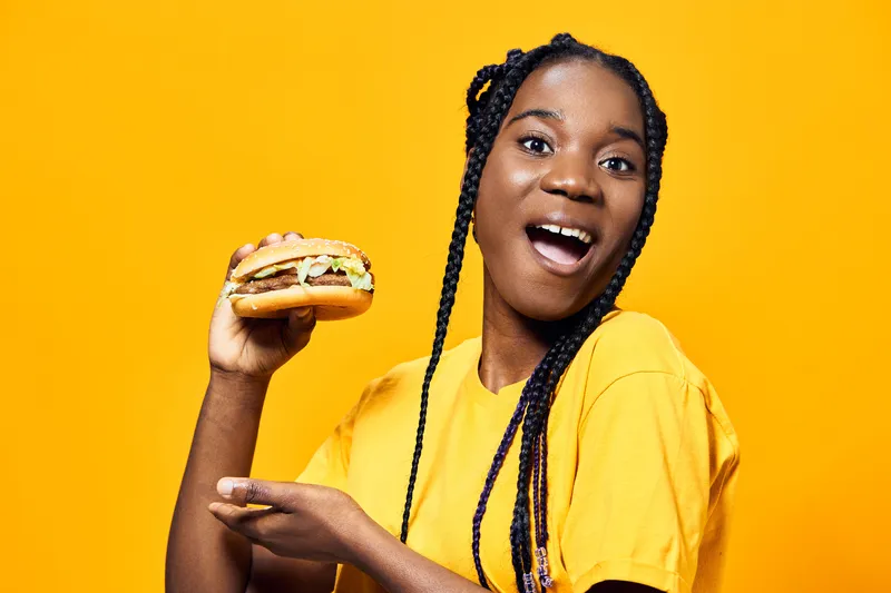 Happy young woman with braids enjoying a delicious burger against a vibrant yellow background, expressing excitement and joy, perfect for food advertising