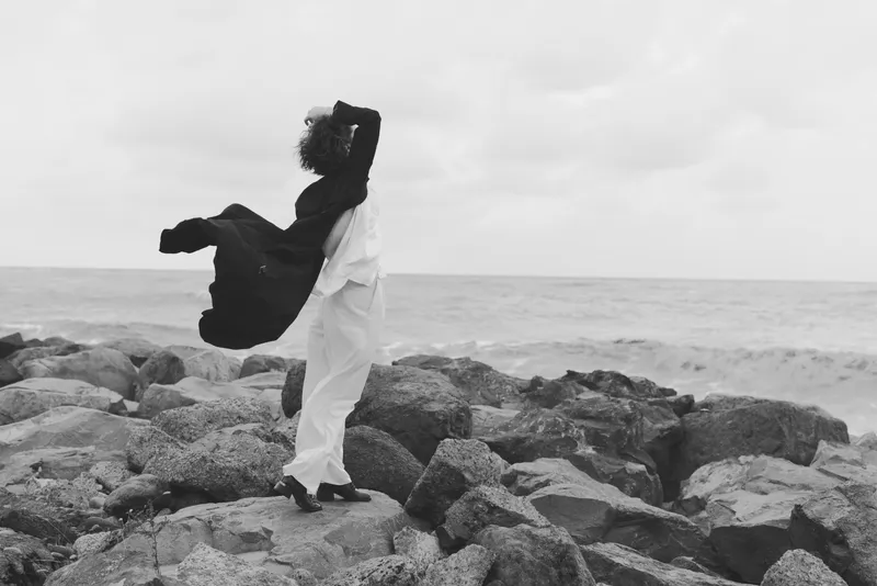 Silhouette, figure, sea, rocks, coast, clothing, standing, outdoors, monochrome scene with wind and horizon showing a bold moment of calm against rugged shoreline