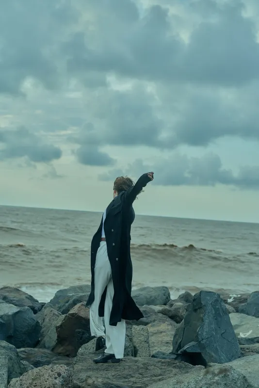 Individual in a long coat on a rocky coast, facing wind and sea, striking a bold pose beneath a dramatic sky, conveying motion and style.