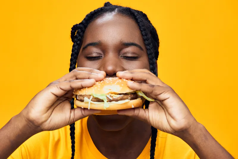 Happy young Black woman enjoying a big hamburger against a vibrant yellow background, radiating joy and excitement for delicious fast food