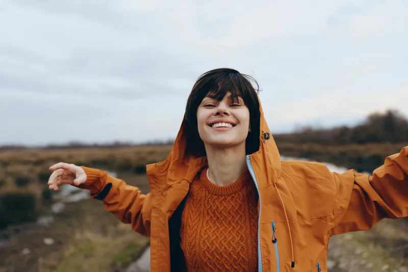 Woman lifestyle outdoor smiling in autumn, orange jacket and knit sweater, stands on countryside trail with arms open, natural portrait, cloudy sky background, cheerful mood today