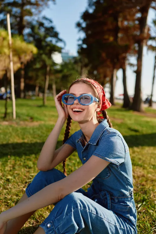 smiling woman wearing blue sunglasses and denim outfit sitting on grass enjoying sunny day outdoors with trees in background and red headband accessory