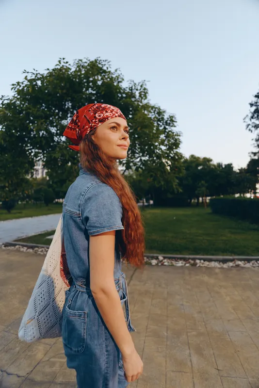 young woman with long hair wearing a red bandana and denim outfit standing outdoors in a park with green trees in warm natural light