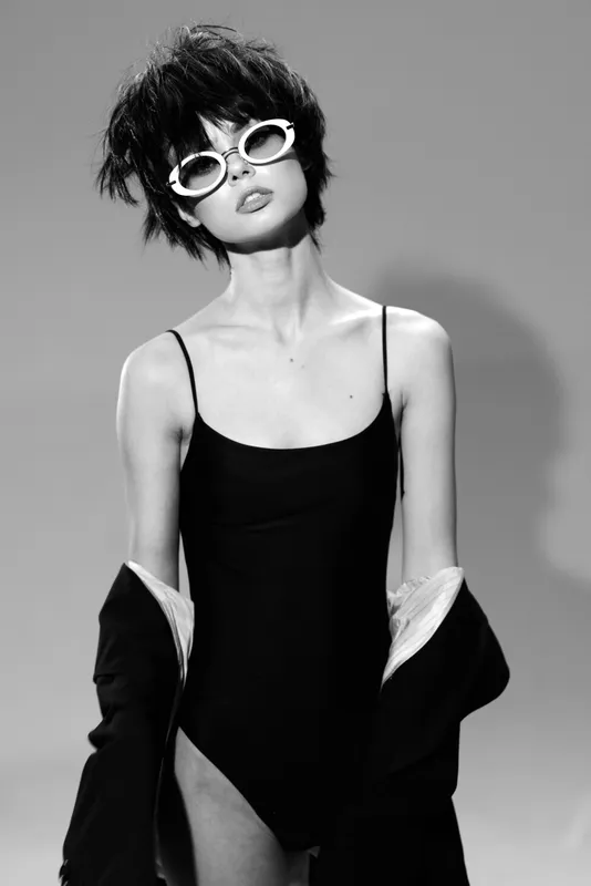 Stylish woman in black swimsuit and white sunglasses posing confidently in black and white minimalistic setting