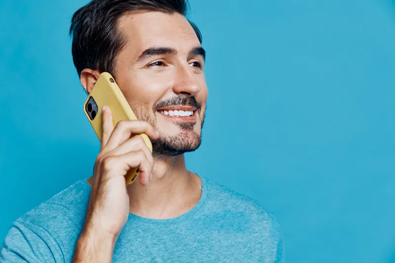 Smiling man talking on mobile phone, wearing light blue shirt, cheerful expression, isolated on bright solid blue background. Modern lifestyle concept.