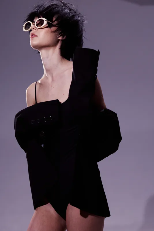 Stylish woman in black outfit wearing sunglasses posing with confident attitude and modern fashion style on grey background