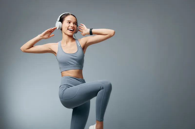 Fitness woman in athletic wear performs a dynamic pose with headphones in a sleek studio, showcasing balance, strength, and rhythm during a workout session.