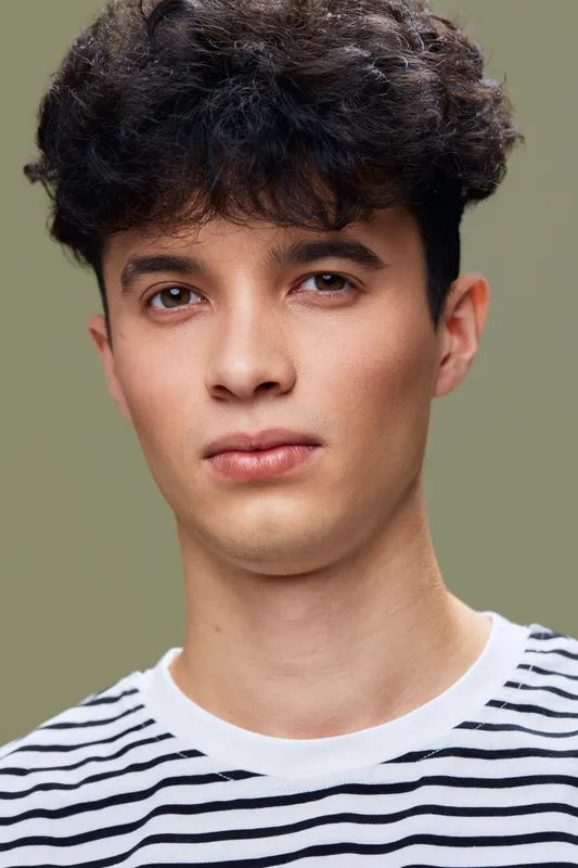 Young man with curly dark hair wearing a striped shirt, looking directly at the camera with a neutral expression against a soft olive background.