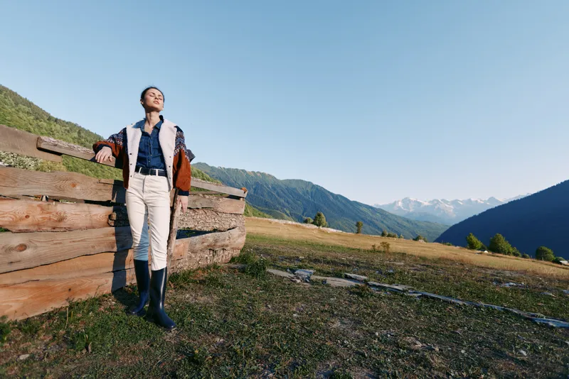 Woman hiking in nature near a wooden fence on a mountain meadow, standing with backpack and boots while enjoying a scenic view and fresh air during a peaceful outdoor adventure.