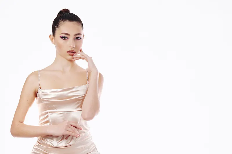 Young woman with high bun hairstyle wearing satin dress poses thoughtfully against white background. Creative beauty fashion concept showcasing elegance and style.