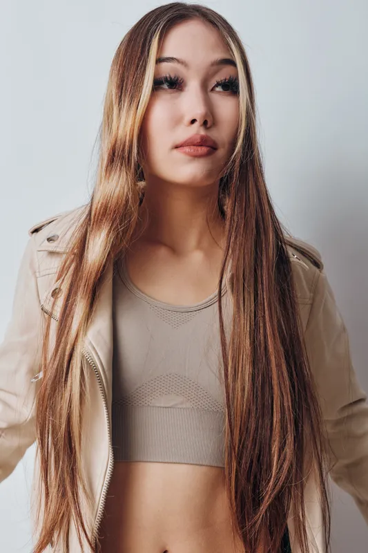 Young woman with long straight hair wearing beige crop top and jacket posing against white wall in studio for fashion portrait and lifestyle concept.