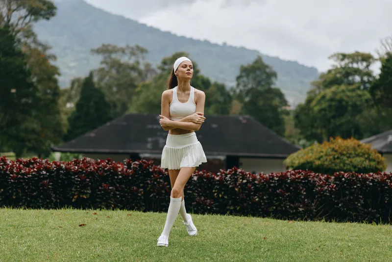 Young athletic woman in a white sports outfit, demonstrating confidence and joy while walking on a green lawn in a beautiful outdoor setting