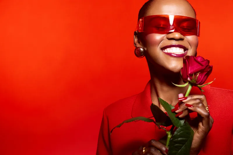 Smiling black woman with dark skin wearing red sunglasses and red outfit holds a red rose on a red background. Concept of beauty, fashion, confidence, and elegance.