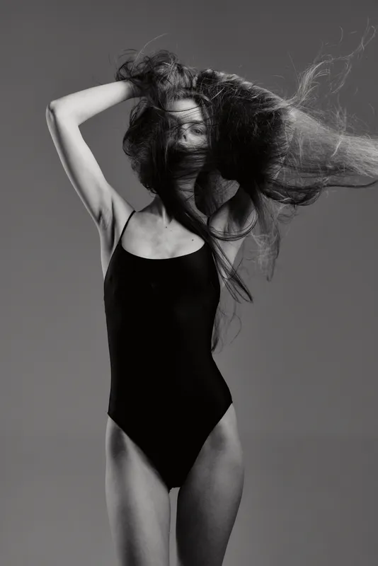 Black and white portrait of a young woman with flowing hair wearing a black bodysuit in a studio setting.