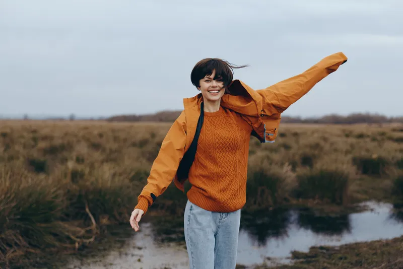 Woman lifestyle outdoors, smiling casual jeans, autumn countryside field near riverbank, walking on wet path, orange sweater jacket, windy hair, evening light, nature travel portrait