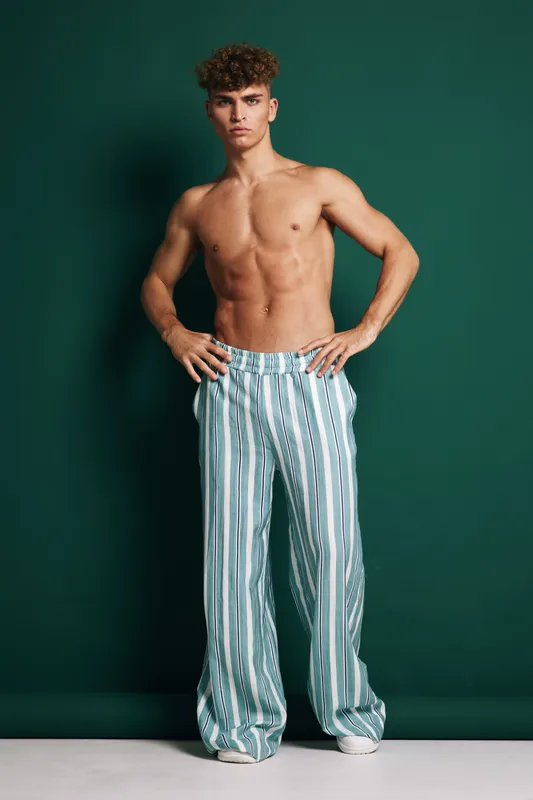 Young man with bare chest standing against teal background wearing loose striped pants with hands on hips, looking confident and posing in fashion studio environment.