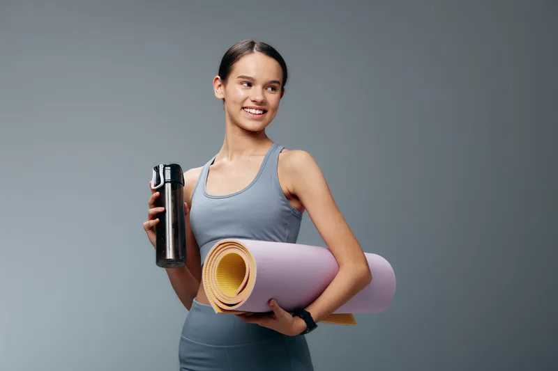 Woman fitness athlete holding a water bottle and yoga mat, ready for a workout. Sporty, focused mood with clear gear and a calm studio backdrop, showcasing an active lifestyle and training readiness.