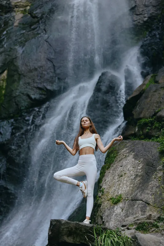 Young woman practicing yoga meditation in white sportswear near a powerful waterfall, surrounded by natural rocks and lush greenery, demonstrating balance and calmness.