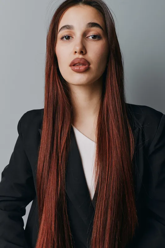 Woman with long straight hair and natural makeup wearing black blazer and white top looking confident and serious in studio with plain background for fashion and beauty portrait.