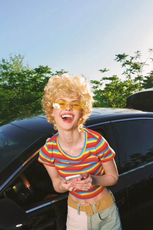 A joyful woman with curly blond hair in a striped shirt and yellow sunglasses laughs beside a sleek car under bright daylight, capturing a carefree retro outdoor lifestyle.
