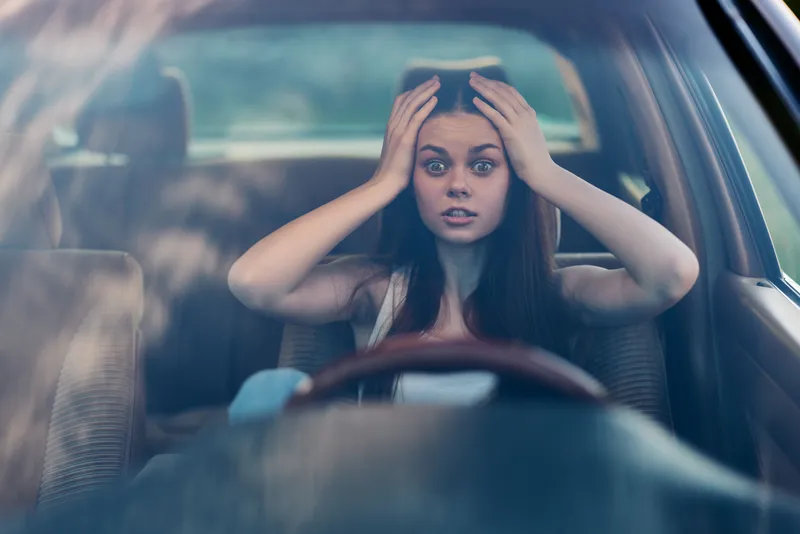 Young woman feeling stressed in car, holding her head in frustration while driving through nature, conveying anxiety and urgency.