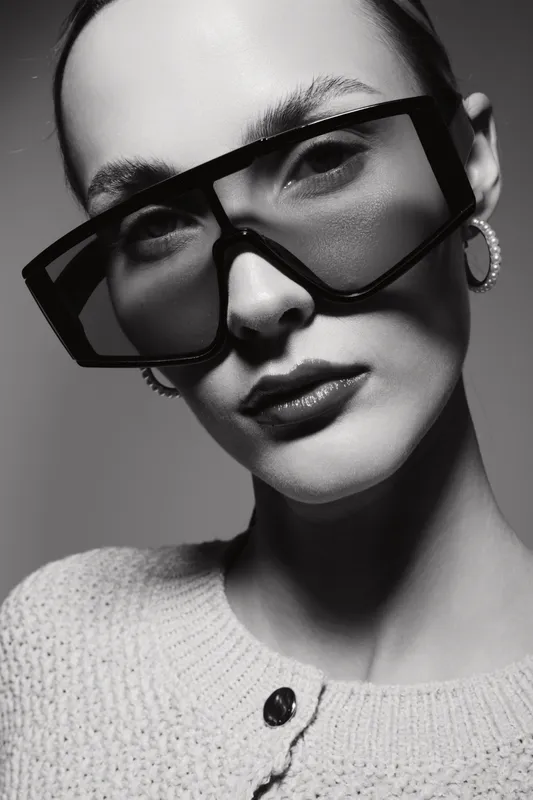 Young white woman with pale skin wearing large black sunglasses and pearl earrings poses in soft studio light. Minimalist fashion portrait with stylish knitwear and confident expression.
