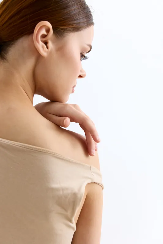 Young woman with bare shoulder in beige top poses gracefully against white background, showcasing soft skin and natural beauty in a minimalistic style.