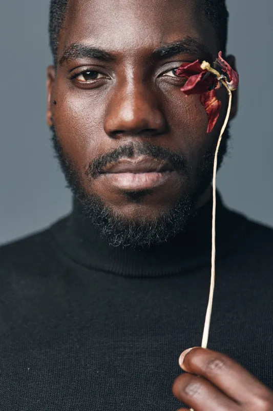 African man with dark skin holding dried flower near face in creative conceptual visual style. Portrait with serious expression and artistic mood on neutral background.