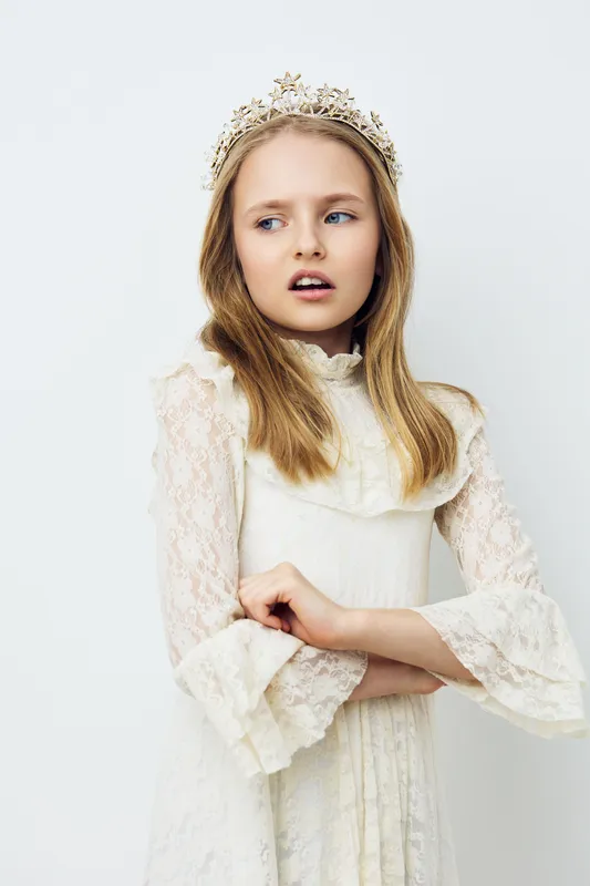 A portrait of a young girl with a crown, wearing a delicate white lace dress and displaying a thoughtful expression against a simple light background