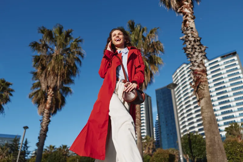 Young woman in a red trench coat walks confidently outdoors on a sunny day with palm trees and modern buildings in the background, smiling and enjoying the moment.