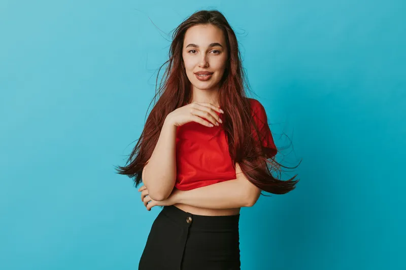 Young woman with long hair wearing red shirt and black pants posing with hand near face on blue background. Casual style, confident look and natural makeup for portrait photography.