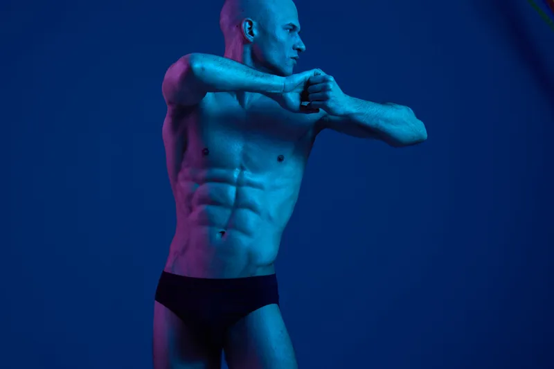 Athletic man posing gracefully in a dramatic light, showcasing toned muscles and strength. The color palette emphasizes deep blue tones, evoking a powerful mood.