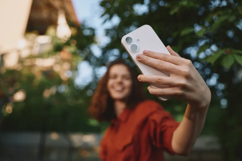 Young woman with brown hair takes a selfie outdoors, smiling softly as she holds a smartphone toward her face with a leafy background, a casual sunlit moment of everyday confidence