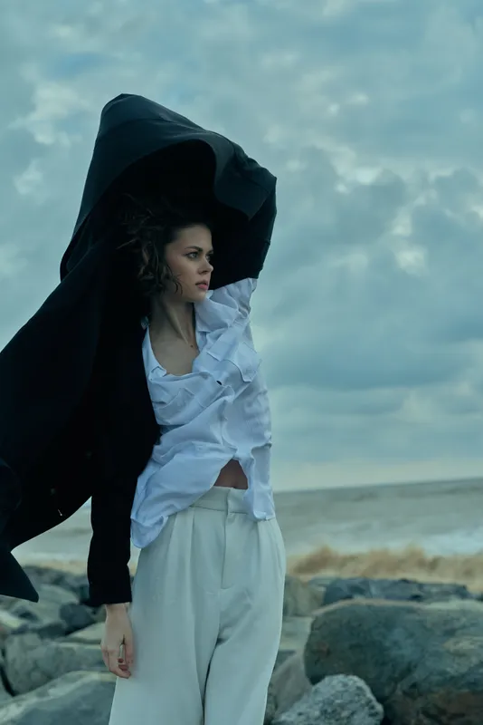 Wind swept coast scene with a hooded cloak, flowing fabric, and a focused expression on a woman by the sea, showcasing dramatic fashion and movement against stormy skies and rocky shoreline