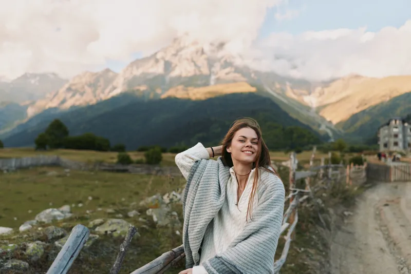 Smiling woman in cozy sweater sitting on wooden fence in rural mountain landscape. Relaxed lifestyle concept with natural light and scenic background.