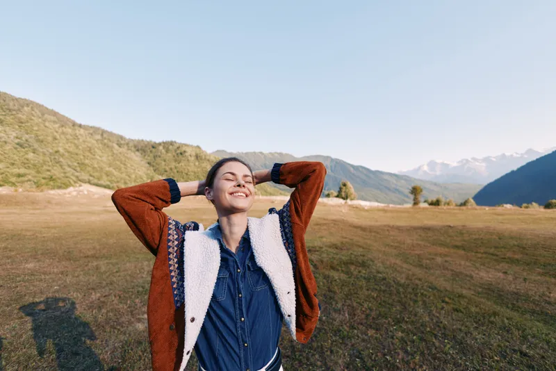 Woman smile in nature with mountains and open field, hands behind head enjoying outdoors and relaxation. Portrait of carefree young adult in casual jacket at sunset.