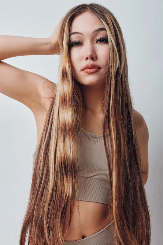 Young woman with long straight hair and casual wear posing with hand behind head against plain background. Portrait of female model with natural makeup and relaxed expression.