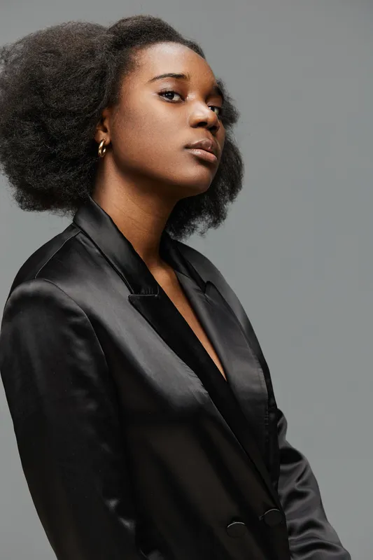 Confident young Black woman in a stylish black satin blazer, exuding elegance and grace against a neutral grey background, perfect for fashion or beauty concepts