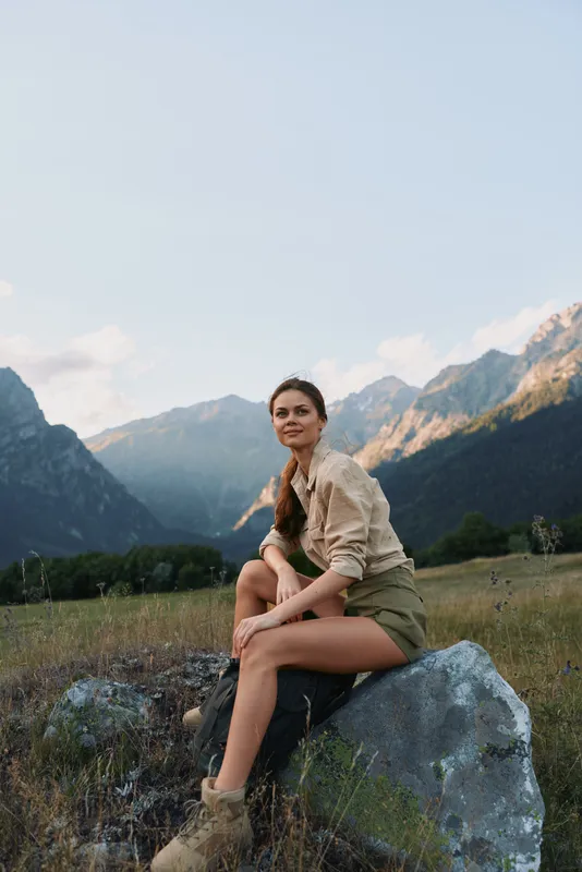 Casual outdoor woman sits on a rock in a mountain valley, wearing a light shirt and shorts, enjoying nature, solo lifestyle moment, serene alpine scenery behind