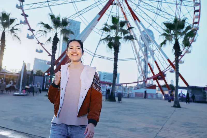 Woman smiles near a large ferris wheel at an outdoor plaza, wearing a brown and white jacket with jeans, capturing a cheerful travel moment and urban amusement atmosphere